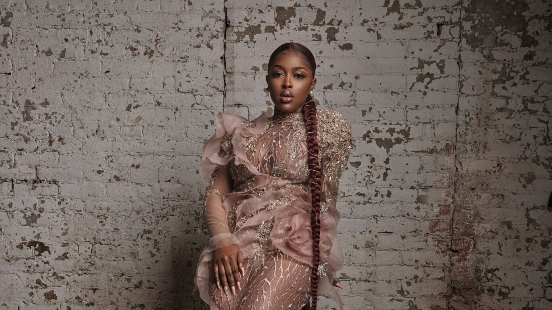 Leila Dey wearing a milk chocolate coloured dress sits on a stool infront of a white brick wall