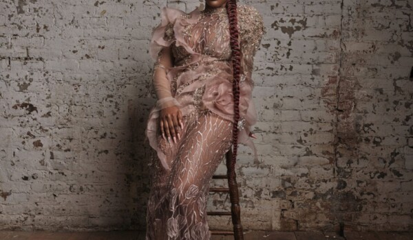 Leila Dey wearing a milk chocolate coloured dress sits on a stool infront of a white brick wall