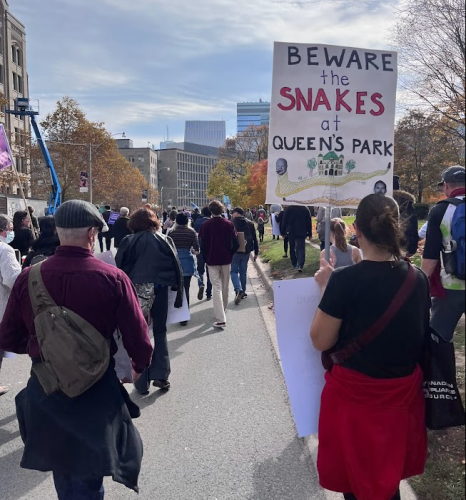 People in a line are walking outside on a road. The person on the right is holding a large white sign on a stick with writing and drawings on it.