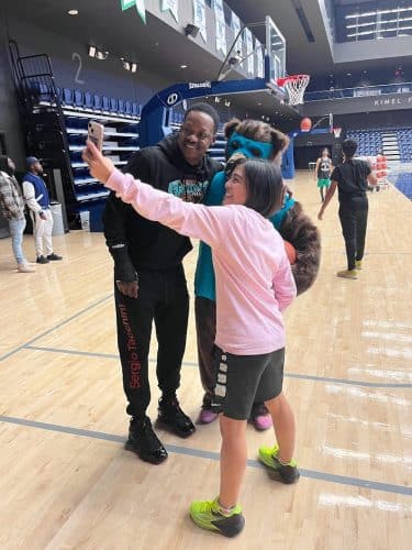 A group of speaking smiling and looking into a phone being held by an arm. There is a basketball net and blue seats in the background.