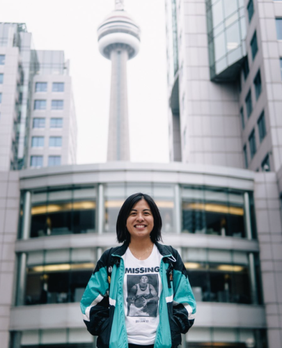 A person smiling and wearing a white shirt with a teal jacket. There are grey buildings or various heights with several windows in the background.