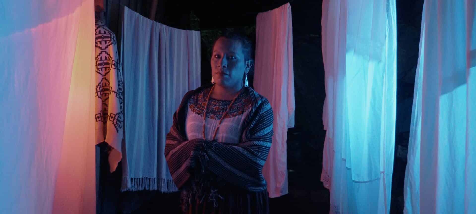 An image of Monica from the Guatemalan short film IXIM: El amor no tiene género. She sits in front of hanging white sheets lit with blue and pink lights.