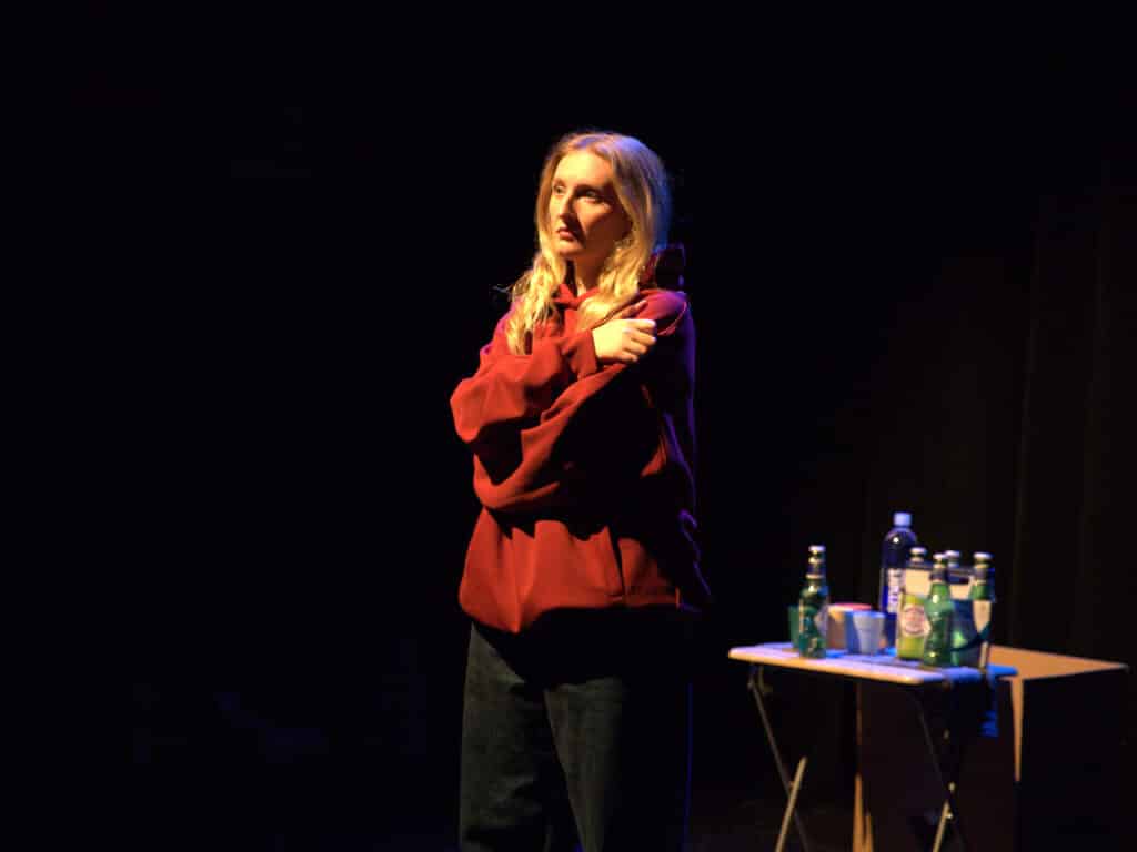 Actress dressed in black jeans, a red hoodie, with a concerned or somewhat exasperated look on her face, in a self hug sort of posture. She is lit by stage light, standing next to a small folding table with 4 small multicolored plastic tumbler glasses, a 6-pack of Peroni beer bottles, and a water bottle. The background is dark, making her a strong focal point amidst the black.