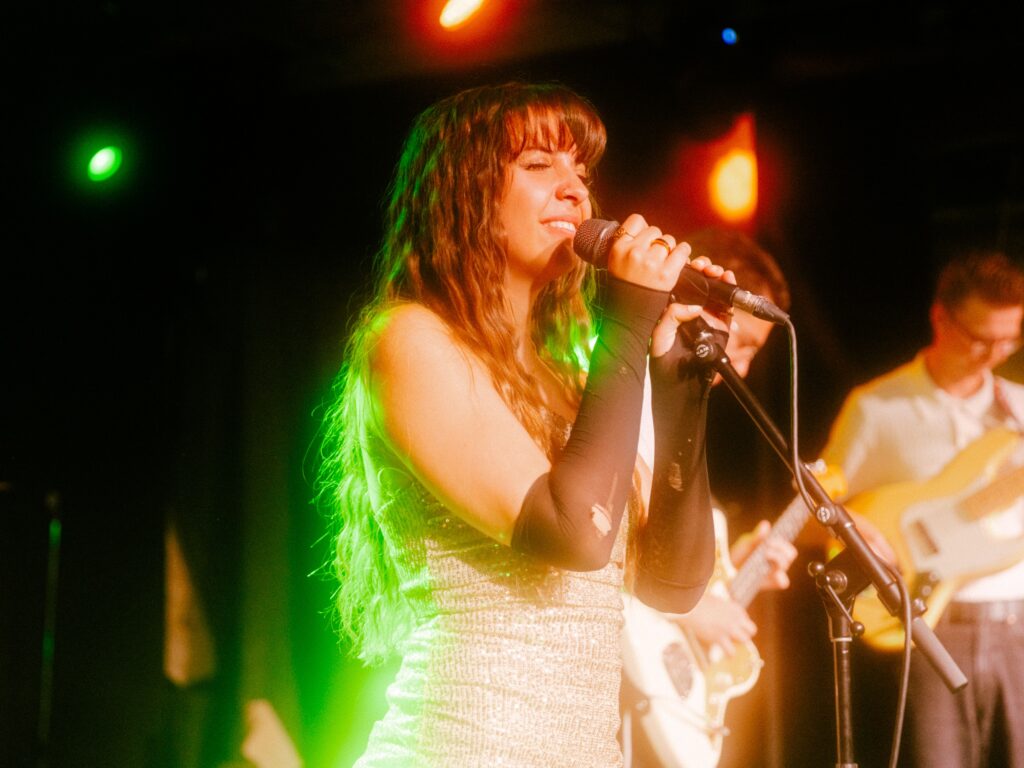 Ella Forrest, in a shimmery silver outfit, with long black fingerless gloves, singing on stage while holding a microphone resting in a stand. A stagelight behind her is illuminating her hair a bright green.