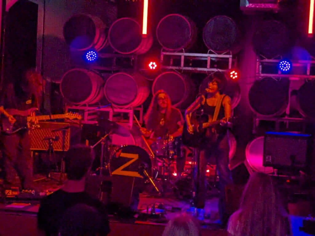 Zoh Amba in bluejeans and a black tank top plays an acoustic guitar, mouth open as she sings into a mic, with electric guitarist hanging his head down and his shaggy hair covering his face. A drummer in the background with long hair and square framed glasses plays along. The stage is in blue and red lights with the round tops of beer kegs lined in rows behind them as the stage backdrop.